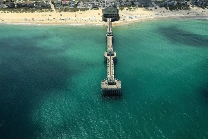 Aerial view of a pier extending into turquoise ocean from a sandy beach with buildings in the background.